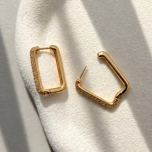 May include: A pair of gold-toned rectangular hoop earrings. Each earring features a hinged post closure and a front-facing edge encrusted with small, clear, sparkling stones. The design is geometric with rounded corners and a polished metal finish.
