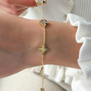 May include: Gold-toned bracelet featuring a series of decorative charms. The charms include a four-petal flower, a diamond-shaped element, a four-point star, and other geometric shapes. The bracelet is worn on a wrist, with a white background.