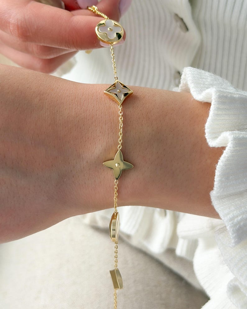 May include: Gold-toned bracelet featuring a series of decorative charms. The charms include a four-petal flower, a diamond-shaped element, a four-point star, and other geometric shapes. The bracelet is worn on a wrist, with a white background.