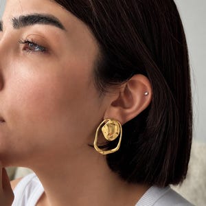May include: A pair of gold-colored earrings. Each earring features an irregular, organic shape with a textured, circular element at the center, surrounded by a smooth, open oval. The earrings are worn on a person's ear.