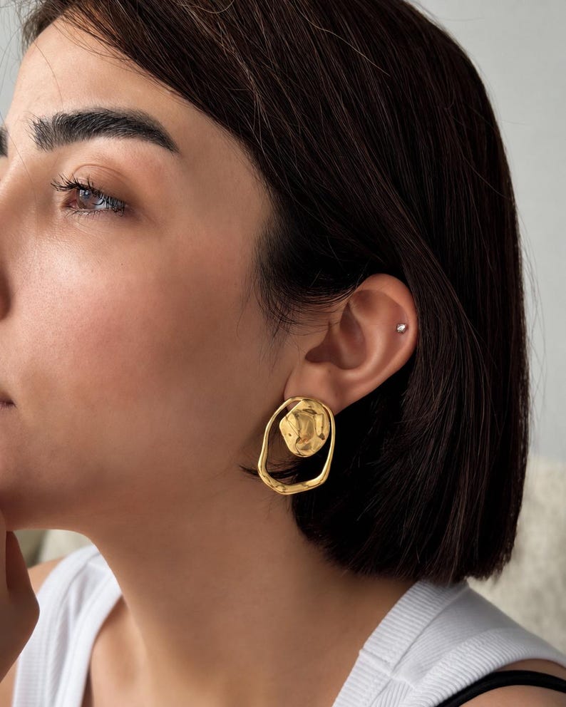 May include: A pair of gold-colored earrings. Each earring features an irregular, organic shape with a textured, circular element at the center, surrounded by a smooth, open oval. The earrings are worn on a person's ear.