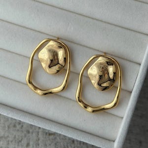 May include: A pair of gold-colored earrings. Each earring features a textured, irregular-shaped gold center piece within a smooth, oval-shaped gold frame. The earrings are displayed on a white, textured surface.