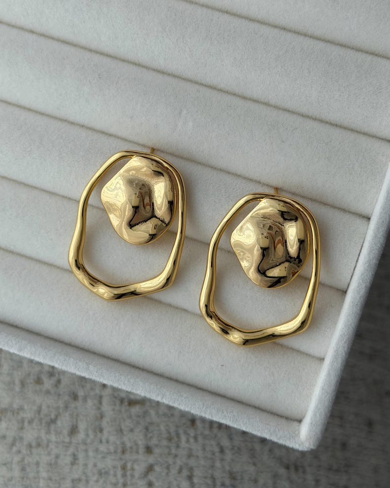 May include: A pair of gold-colored earrings. Each earring features a textured, irregular-shaped gold center piece within a smooth, oval-shaped gold frame. The earrings are displayed on a white, textured surface.