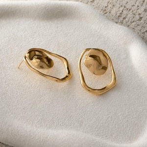 May include: A pair of gold-colored earrings. Each earring features an irregular, open oval frame with a textured, rounded element inside. The earrings are displayed on a white, textured surface.