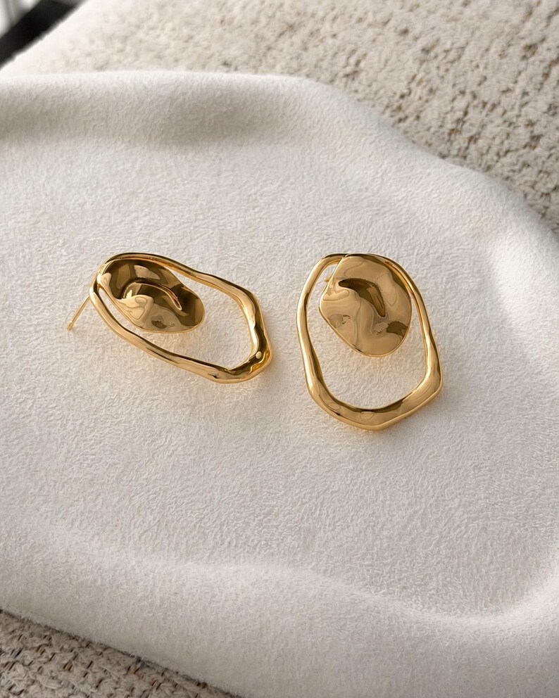 May include: A pair of gold-colored earrings. Each earring features an irregular, open oval frame with a textured, rounded element inside. The earrings are displayed on a white, textured surface.