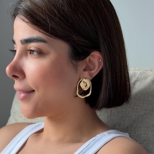 May include: Gold-toned, abstract-shaped earrings. The earrings feature a textured, circular design within an open, irregular oval. The jewelry is worn on the ear. The earrings are a bright, shiny gold color.