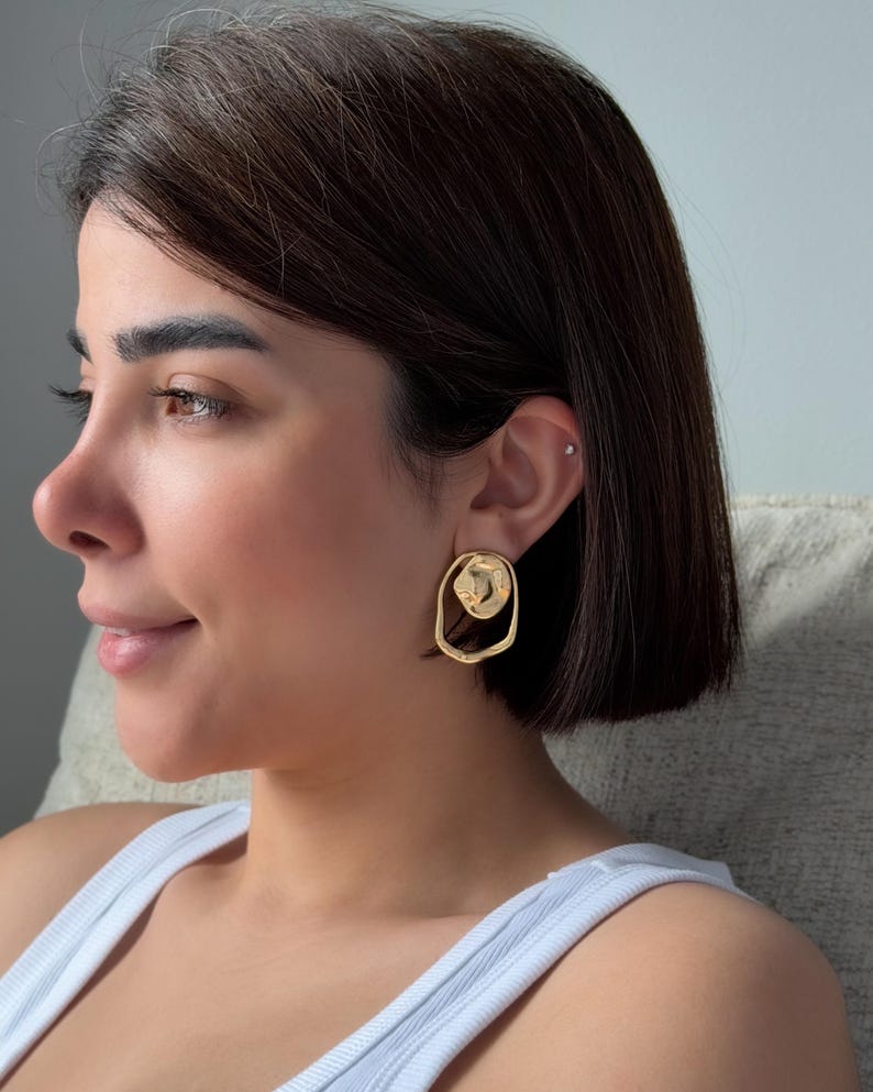 May include: Gold-toned, abstract-shaped earrings. The earrings feature a textured, circular design within an open, irregular oval. The jewelry is worn on the ear. The earrings are a bright, shiny gold color.
