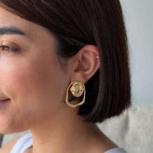 May include: Gold-toned, abstract-shaped earrings. The earrings feature a textured, irregular design with a circular element within an oval frame. The earrings are a bright, shiny gold color.