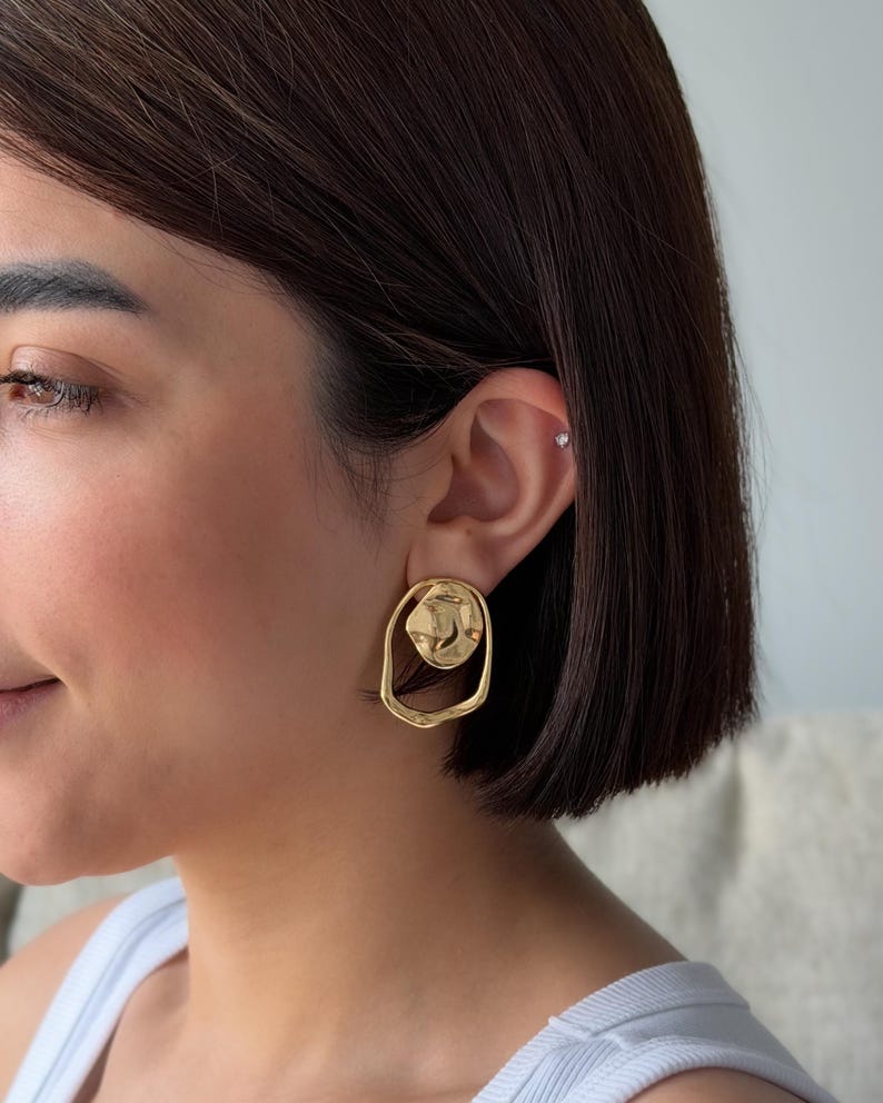 May include: Gold-toned, abstract-shaped earrings. The earrings feature a textured, irregular design with a circular element within an oval frame. The earrings are a bright, shiny gold color.