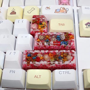 May include: A close-up of a white keyboard with custom keycaps. The keycaps feature various designs, including cartoon images and resin-filled keycaps with miniature sea creatures and red and white accents. The "ALT" and "TAB" keys are visible.