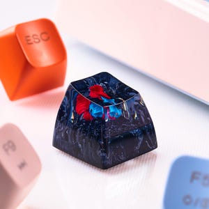 May include: A custom artisan keycap with a dark blue resin base. The keycap features a red and blue design inside. An orange ESC keycap and other keyboard keys are visible in the background.