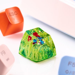 May include: A custom artisan keycap with a vibrant green, yellow, red, and blue design. The keycap has a clear, resin-like appearance, with a miniature landscape inside. Other keyboard keys are visible in the background.