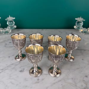 Pode incluir: Seis cálices de prata ornamentados com intrincados desenhos florais e em forma de coração. O interior de cada cálice é dourado. Estão dispostos sobre uma superfície de mármore, mostrando o seu artesanato detalhado.