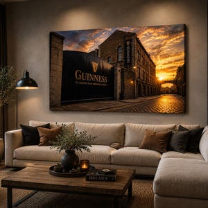 May include: A large canvas print of the Guinness brewery entrance in Dublin, Ireland, at sunset. The image features the brewery's gate with the Guinness logo and text, a cobblestone street, and a vibrant orange and yellow sky. The print is displayed above a beige sectional sofa.