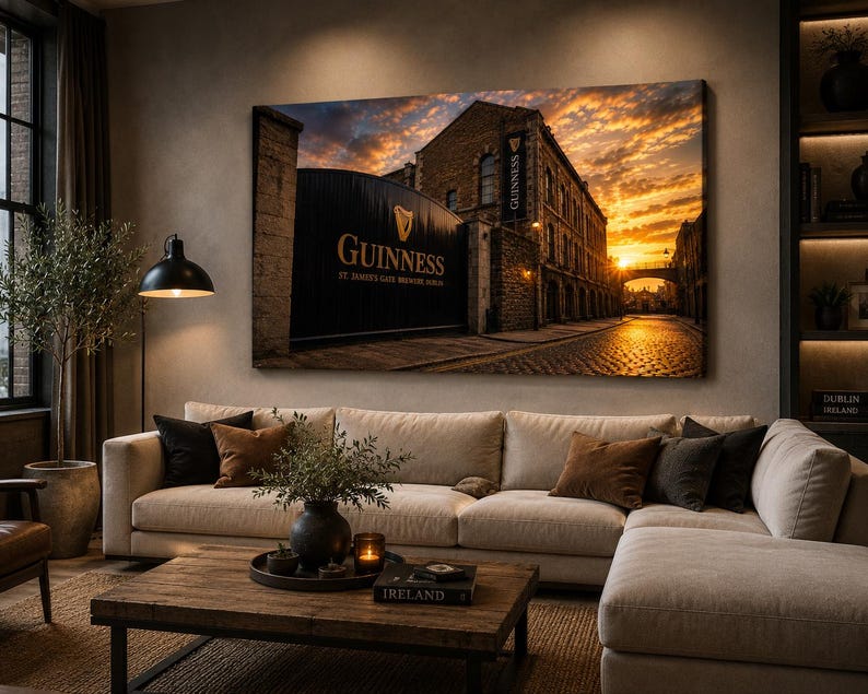 May include: A large canvas print of the Guinness brewery entrance in Dublin, Ireland, at sunset. The image features the brewery's gate with the Guinness logo and text, a cobblestone street, and a vibrant orange and yellow sky. The print is displayed above a beige sectional sofa.