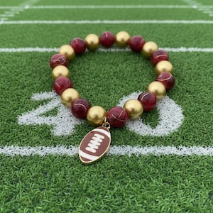 Op de afbeelding: Een armband met bordeauxrode en gouden kralen, versierd met een gouden voetbalbedel. De armband ligt op een groen voetbalveld. De kralen hebben een diameter van ongeveer 0,8 cm.