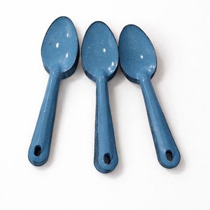 Puede incluir: Tres cucharas de esmalte azul con acabado moteado. Cada cuchara tiene un mango largo y un pequeño agujero en el extremo. Las cucharas están dispuestas sobre un fondo blanco.