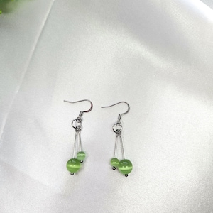 May include: A pair of dangle earrings featuring silver-tone hooks and delicate chains. Each earring has two green, spherical beads, one larger than the other. The earrings are set against a white, silky background.