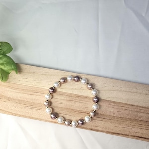May include: A beaded bracelet featuring a mix of white, gold, and dark purple pearls. The bracelet is displayed on a wooden surface against a white background. The pearls are strung together with a gold-colored string.