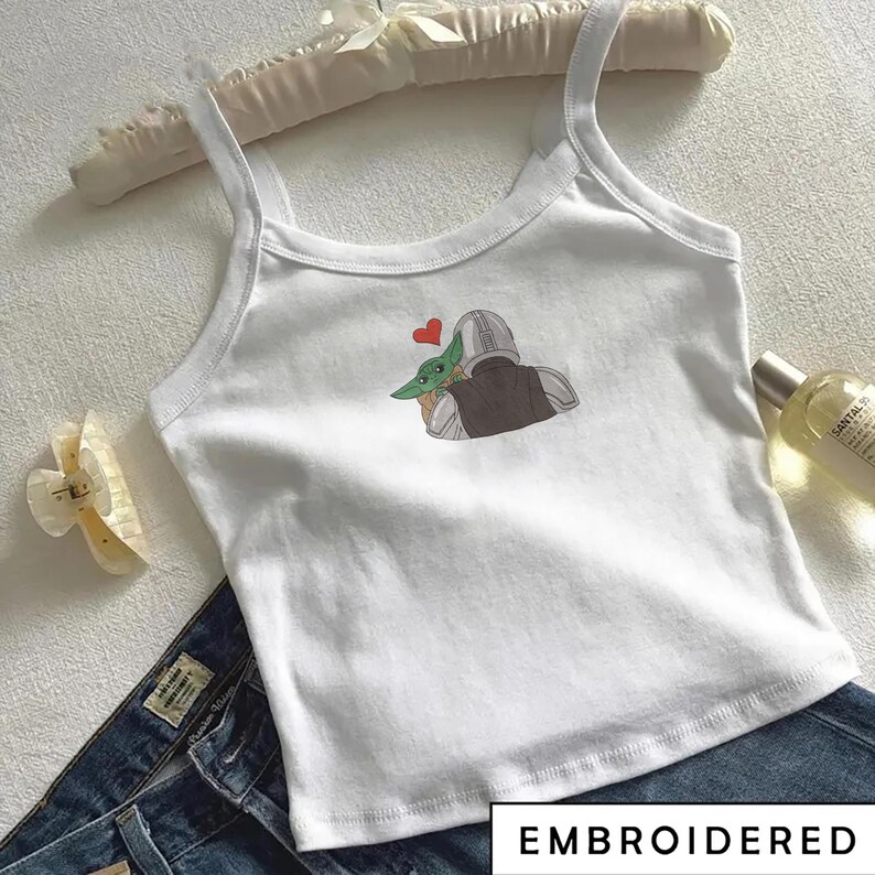 May include: White tank top with a graphic of a character embracing another character with a red heart above them. The word "EMBROIDERED" is printed in a white box at the bottom. The tank top is paired with blue jeans.