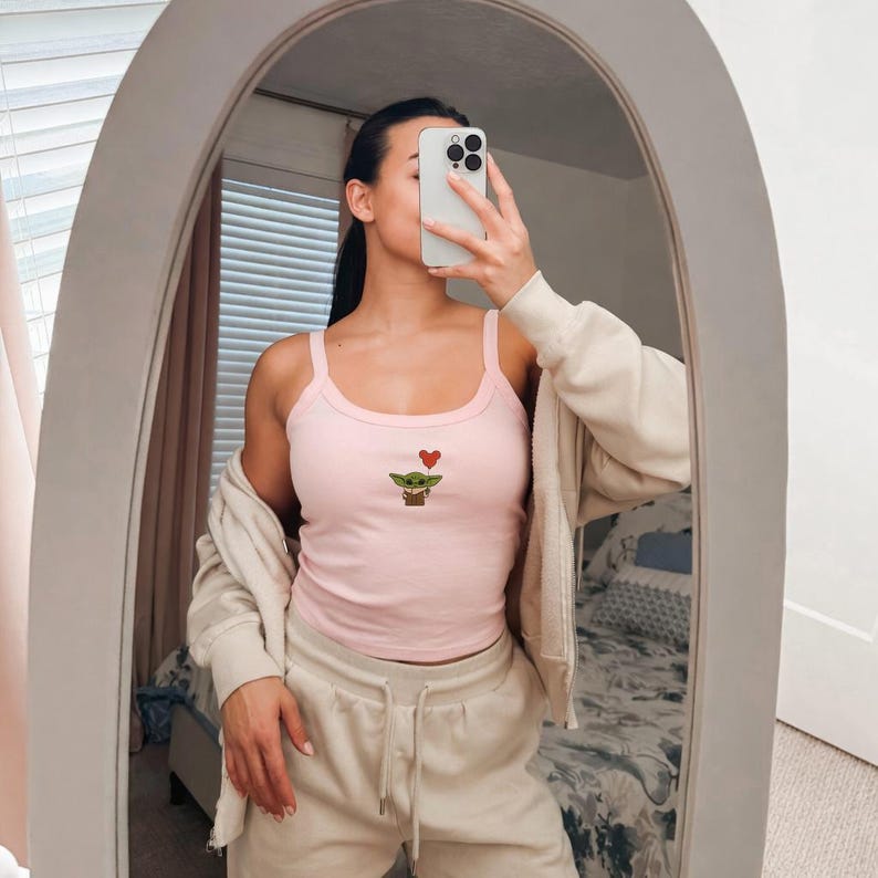 Puede incluir: Una camiseta sin mangas rosa claro con una peque&ntilde;a imagen bordada de un personaje verde y un coraz&oacute;n rojo. La camiseta sin mangas tiene tirantes finos y se usa con una chaqueta beige y un pantal&oacute;n de ch&aacute;ndal.