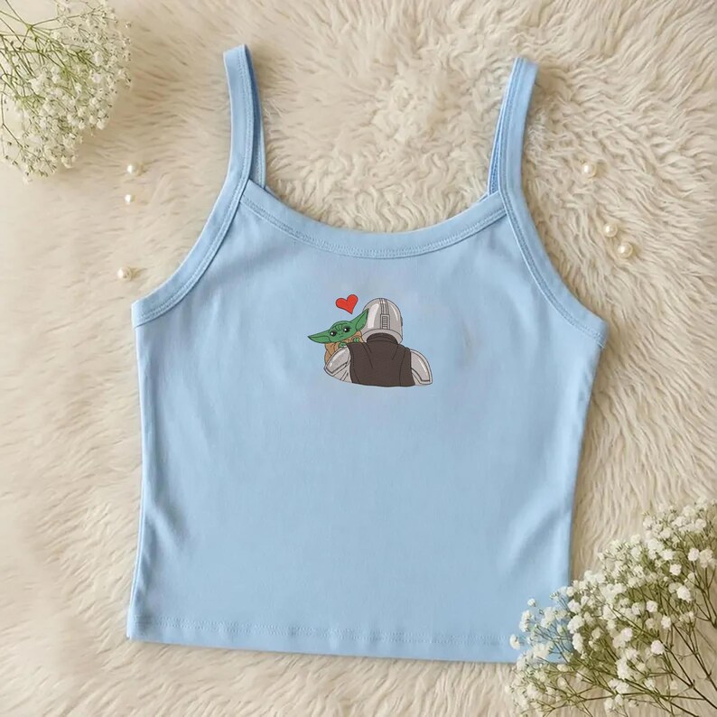 May include: Light blue camisole top with spaghetti straps. The top features an embroidered design of a character holding a green alien with a red heart above them. The top is made of a soft material.