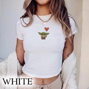 Puede incluir: Top corto blanco con una ilustraci&oacute;n de dibujos animados de un personaje alien&iacute;gena verde sosteniendo un globo rojo en forma de coraz&oacute;n. La palabra "WHITE" est&aacute; impresa en letras negras grandes y en negrita en la parte inferior de la camisa.