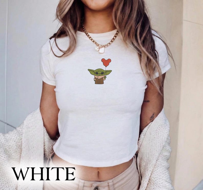Puede incluir: Top corto blanco con una ilustraci&oacute;n de dibujos animados de un personaje alien&iacute;gena verde sosteniendo un globo rojo en forma de coraz&oacute;n. La palabra "WHITE" est&aacute; impresa en letras negras grandes y en negrita en la parte inferior de la camisa.