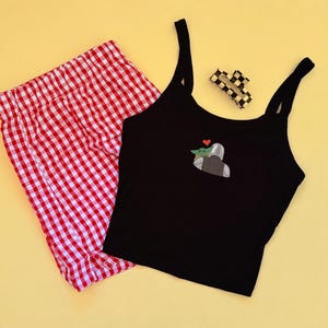 May include: A black tank top with a graphic of a character and a red heart, paired with red and white checkered shorts. A checkered hair clip rests nearby. The items are arranged on a yellow background.