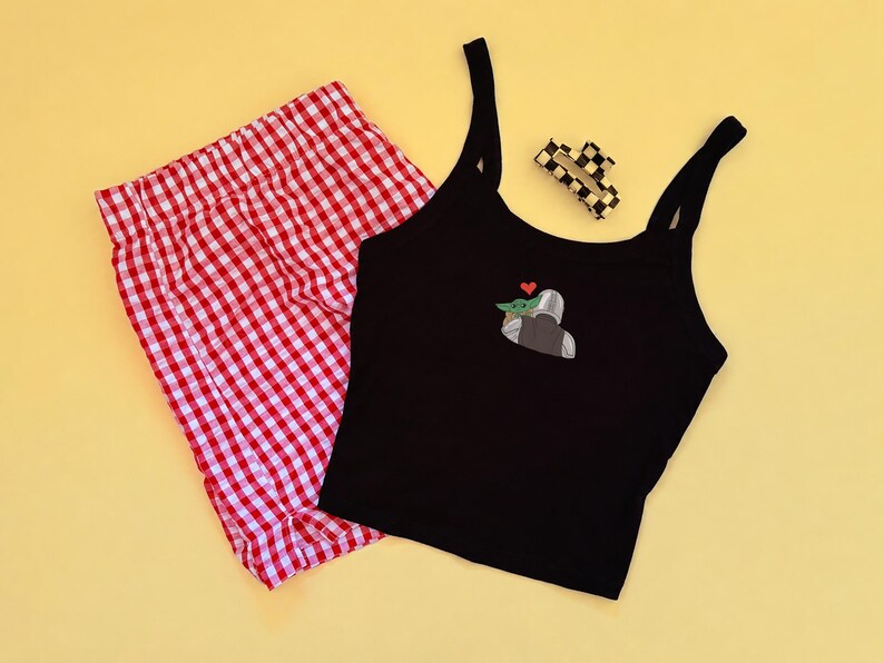 May include: A black tank top with a graphic of a character and a red heart, paired with red and white checkered shorts. A checkered hair clip rests nearby. The items are arranged on a yellow background.