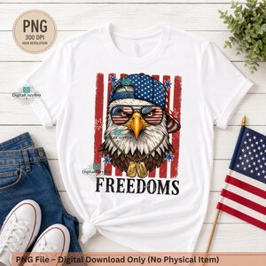 Puede incluir: Camiseta blanca con un diseño de águila patriótica. El águila lleva gafas de sol con un diseño de bandera estadounidense y una gorra azul con estrellas blancas. La palabra "FREEDOMS" está impresa debajo. La camiseta tiene cuello redondo.