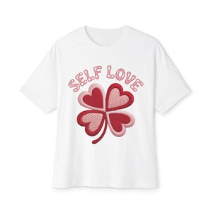 Peut inclure: T-shirt blanc avec un motif de trèfle composé de quatre cœurs roses et rouges. Les mots "SELF LOVE" sont brodés au-dessus du trèfle. Le t-shirt a des manches courtes et un col rond.
