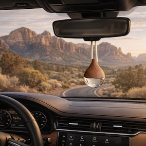 May include: A car interior with a hanging air freshener. The air freshener has a wooden top and a clear glass bottom. The car's dashboard and steering wheel are visible, with a scenic mountain view in the background.