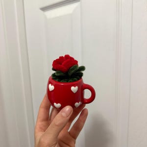 May include: A small, red ceramic teacup decorated with white hearts, holding a red rose. The teacup has a handle and is filled with green foliage. The image is set against a white background.