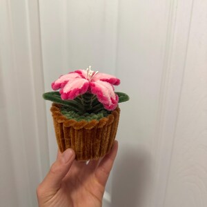 May include: A decorative flower in a cupcake-shaped container. The flower has pink petals with a darker pink trim, green leaves, and white stamens. The container is made of a textured, brown material. The item is handmade.