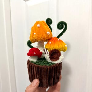 May include: A whimsical, handmade cupcake decoration featuring three colorful mushrooms. The mushrooms have orange, red, and yellow caps with white pearl accents. The cupcake base is brown with a green grassy top.