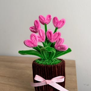 Può includere: Composizione floreale decorativa con fiori rosa a forma di cuore, steli e foglie verdi, in un vaso marrone a coste, legato con un nastro rosa. Un affascinante elemento decorativo.