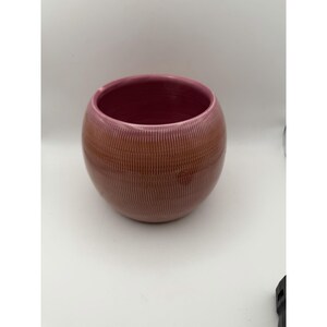 May include: A round ceramic bowl with a smooth, deep pink interior and a textured exterior featuring vertical, etched lines. The outer surface displays a gradient of warm, earthy brown tones that transition into a soft pink hue near the rim.
