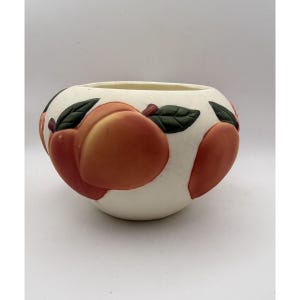May include: A round, off-white ceramic bowl featuring a raised, hand-painted pattern of orange and red peaches with dark green leaves. The bowl has a smooth, matte finish and a wide, open top, showcasing a repeating fruit motif around the exterior.