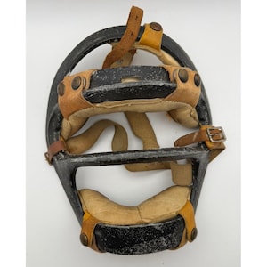 Peut inclure: Un masque de receveur de baseball vintage doté d'une armature en métal noir présentant des signes d'usure et des éclats de peinture. L'intérieur est doublé de coussinets en cuir beige fixés par des lanières en cuir marron et des rivets.