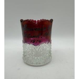 Peut inclure: Un porte-cure-dents en verre avec un bord festonné, présentant une section supérieure teintée en rouge profond et une section inférieure transparente et texturée avec un motif pressé. Le texte Souvenir of Ashland, Ohio 1974 est imprimé en noir.