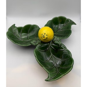 Puede incluir: Una fuente de cerámica con forma de tres hojas verdes grandes, con una pieza central en forma de limón amarillo. La fuente tiene un acabado brillante y probablemente se utiliza para servir aperitivos o con fines decorativos.