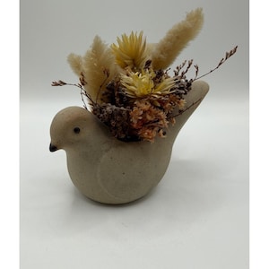 May include: A ceramic bird-shaped planter in a neutral beige color, filled with dried flowers and grasses. The arrangement includes fluffy beige, yellow, and brown elements, creating a natural, textured display. The bird has a small, dark eye and a simple, elegant design.