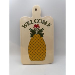 May include: A cream-coloured ceramic wall plaque shaped like a cutting board with a hole at the top. It features the word WELCOME in dark green letters above a yellow pineapple graphic with green leaves and a small red heart at the very top.