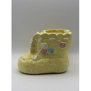 Puede incluir: Una maceta de cerámica amarilla clara con forma de bota. La maceta presenta un diseño con cordones con cordones blancos y ojales dorados. Está decorada con patrones florales en relieve y pequeños acentos florales de colores.