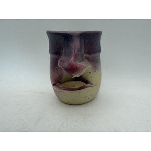 May include: A ceramic mug featuring a sculpted human face on the front. The upper portion of the mug is a deep purple, transitioning into a speckled cream color at the base. The nose and lips are molded in relief, creating a three-dimensional effect.