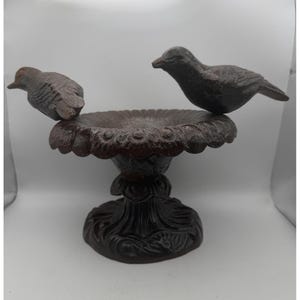 Puede incluir: Un baño para pájaros de hierro fundido en color marrón oscuro con un cuenco circular de borde festoneado, apoyado sobre una base de pedestal decorativa. Dos figuras de aves esculpidas están fijadas en lados opuestos del borde.