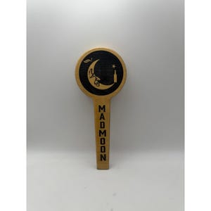 May include: A wooden tap handle with a circular top featuring a black crescent moon design with a bottle and star. The handle has the word "MADMOON" vertically engraved in black. The tap handle is made of light-colored wood.