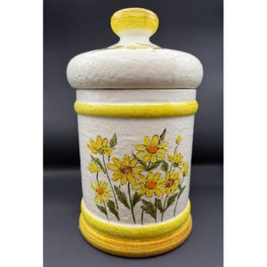 May include: A white ceramic canister with a lid, adorned with yellow daisies and green leaves. The canister has yellow bands at the top and bottom, and a yellow knob on the lid. The design is a charming, vintage-inspired floral pattern.
