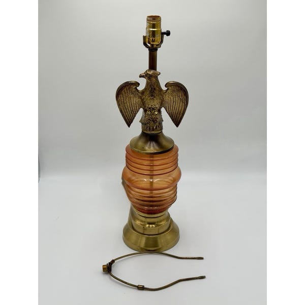 Peut inclure: Une lampe de table avec une figurine d'aigle en laiton montée sur une base en verre ambré nervuré. La lampe comprend une douille en laiton au sommet et une lyre en métal séparée posée devant la base sur une surface blanche unie.